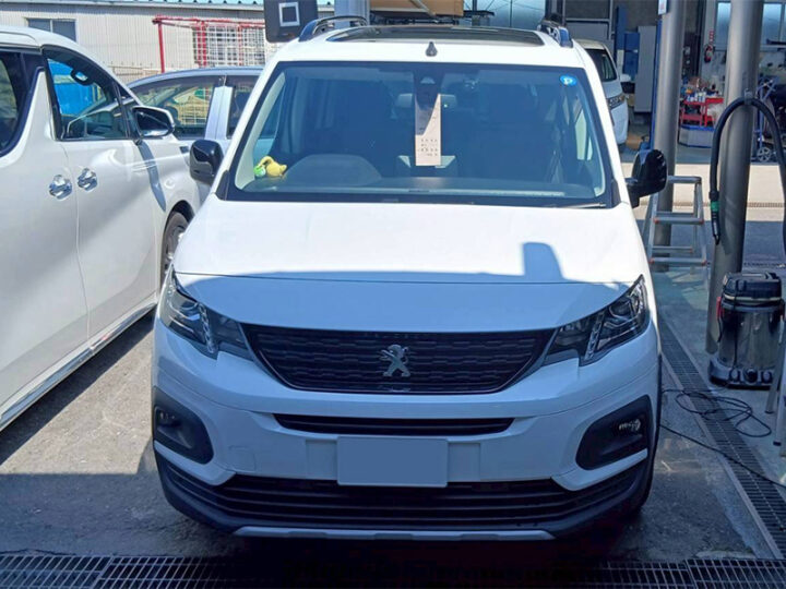 Front view of a white Peugeot van in a service bay, with workshop equipment in the background and another white car beside it