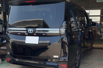 Black Toyota Voxy minivan in a workshop viewed from the rear, with the license plate blurred and workshop tools in the background