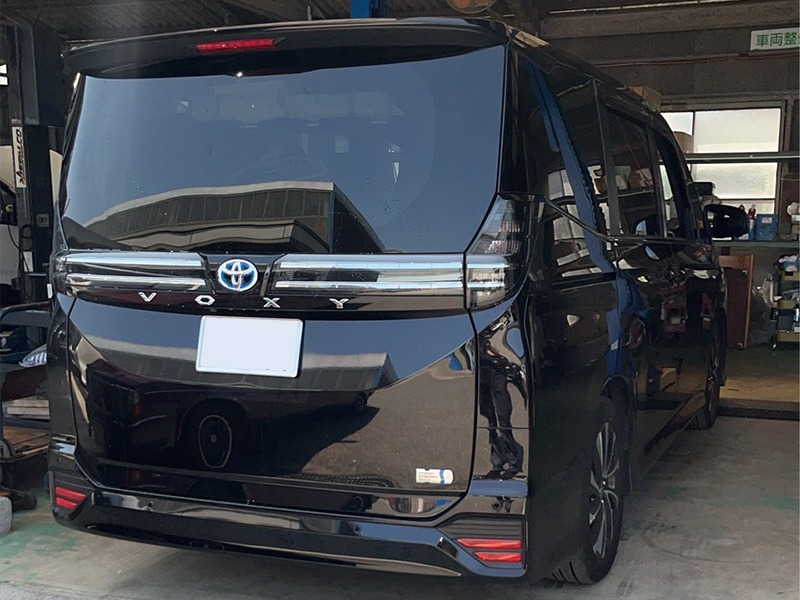 Black Toyota Voxy minivan in a workshop viewed from the rear, with the license plate blurred and workshop tools in the background