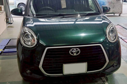 Front view of a dark green Toyota car inside a workshop; round headlights, prominent hexagonal grille with chrome trim, and a blank license plate area.