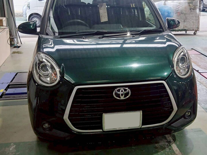 Front view of a dark green Toyota car inside a workshop; round headlights, prominent hexagonal grille with chrome trim, and a blank license plate area.