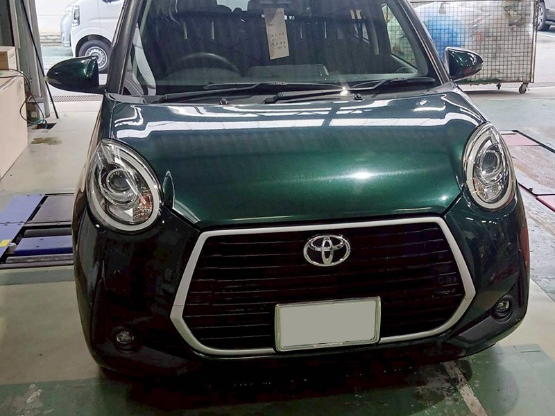Front view of a dark green Toyota car inside a workshop; round headlights, prominent hexagonal grille with chrome trim, and a blank license plate area.
