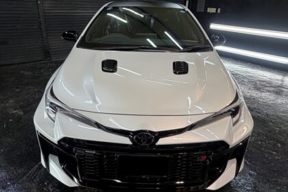 Front view of a white Toyota car facing the camera, with a black grille, dual hood vents, and glossy bumper in a dark garage backdrop.
