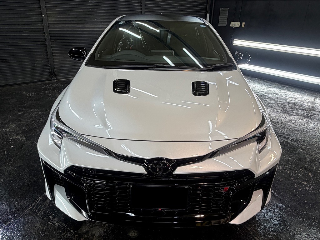 Front view of a white Toyota car facing the camera, with a black grille, dual hood vents, and glossy bumper in a dark garage backdrop.
